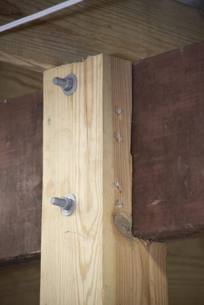 Close-up of a wooden beam joint secured with large bolts and washers, attached to a darker horizontal beam. Several small holes are visible in the lighter wood near the bolts.
