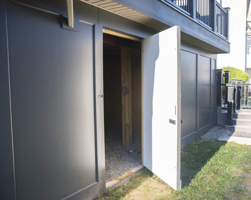 A white door stands open, revealing a dark storage area beneath a raised deck; sunlight casts shadows on the exterior gray walls and nearby green grass.