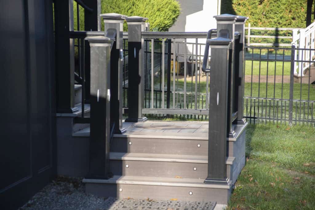 Small outdoor staircase with three steps and black railings, leading up to a gated porch area. The steps are partly in sunlight, and a grassy yard with fences is visible in the background.
