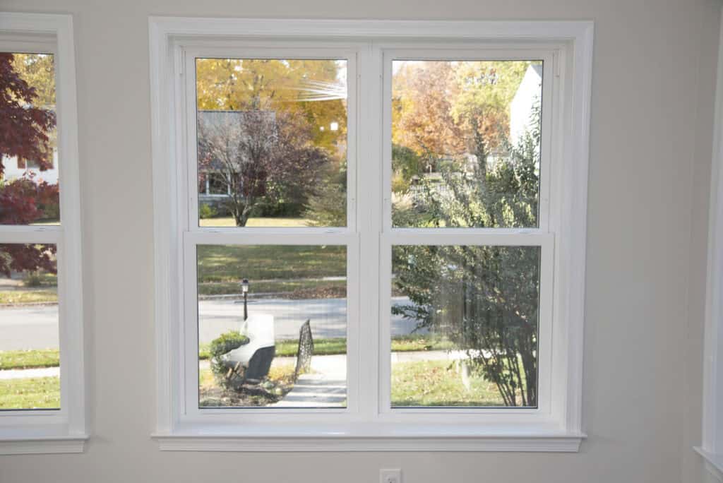 A large double window with white trim overlooks a suburban street lined with trees displaying autumn foliage in shades of yellow, orange, and red. Sunlight brightens the scene outside.