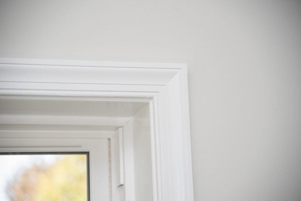Close-up of a white-painted window frame in a light-colored room, showing the upper corner with part of the windowpane and blurred outdoor scenery visible through the glass.