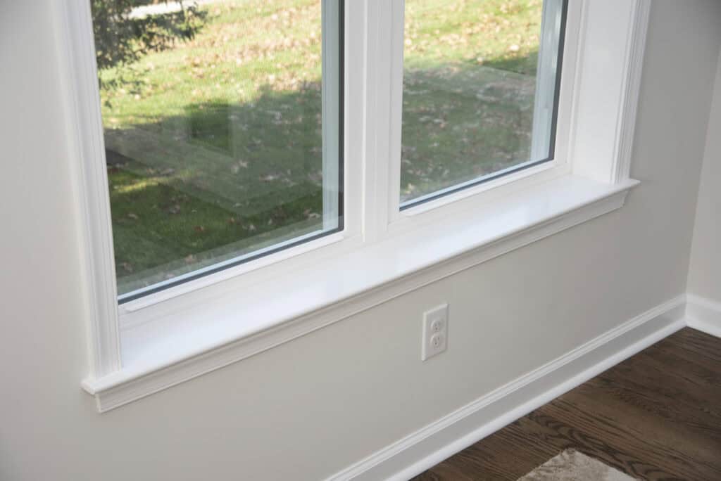 A close-up of a white windowsill beneath a double window, with an electrical outlet on the wall below. Outside, grass and scattered fallen leaves are visible. Part of a wooden floor and a rug can be seen inside.