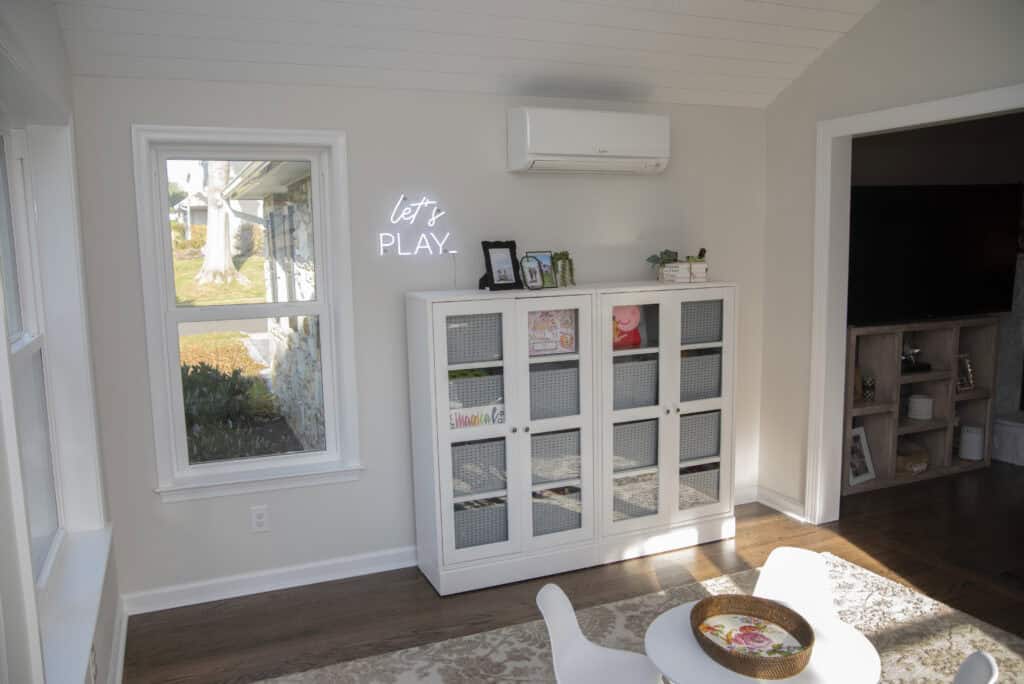 A bright playroom features a white cabinet with mesh doors holding books and toys, a small round table with chairs, and a neon sign on the wall reading lets PLAY. Sunlight streams through the windows.