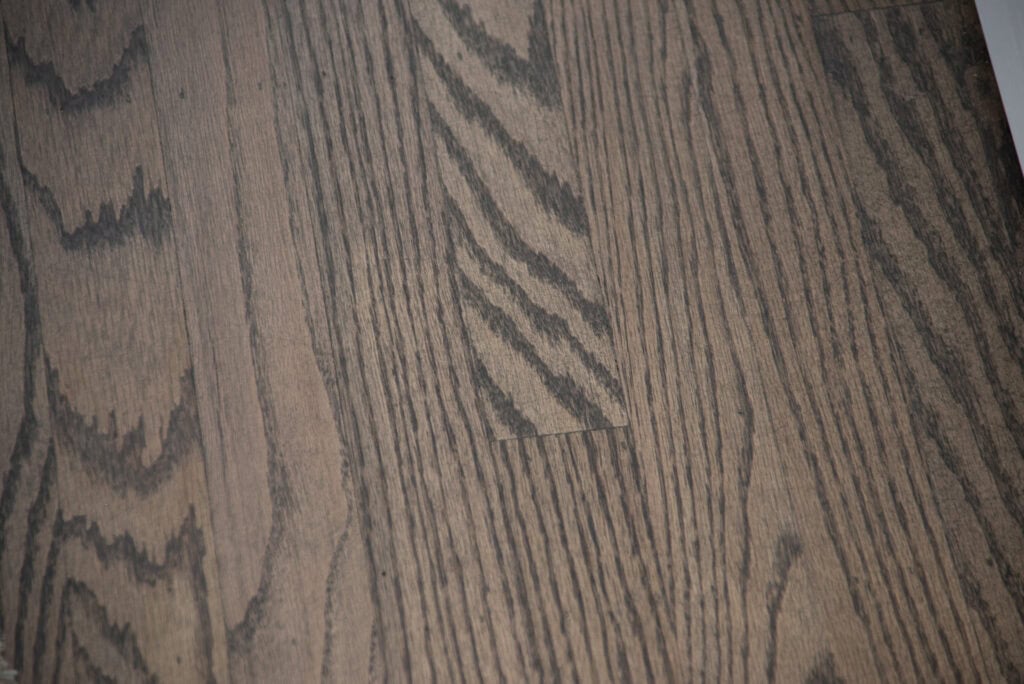Close-up view of a wooden floor with a natural grain pattern in brown tones. The planks have visible lines and textures, giving a rustic and warm appearance.