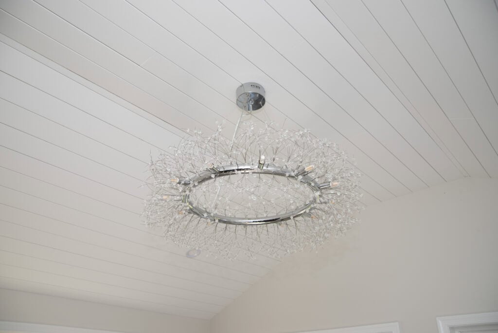 A modern chandelier with a circular metal frame and delicate, branch-like details hangs from a white, wood-paneled ceiling in a bright room.
