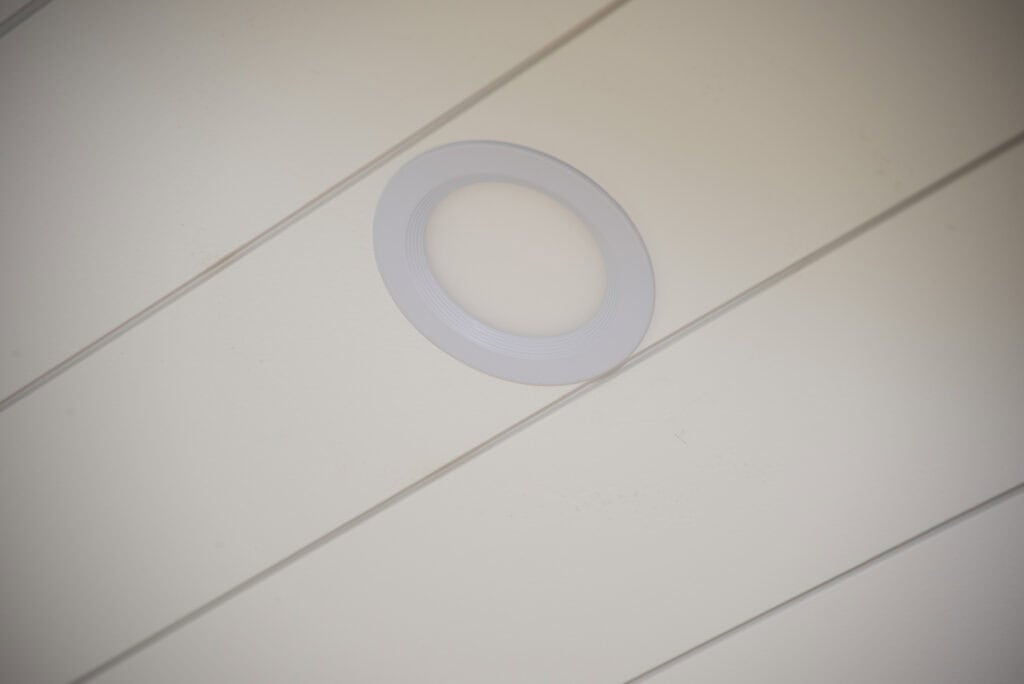 A round, white recessed ceiling light is installed in a beige ceiling with diagonal panel lines.
