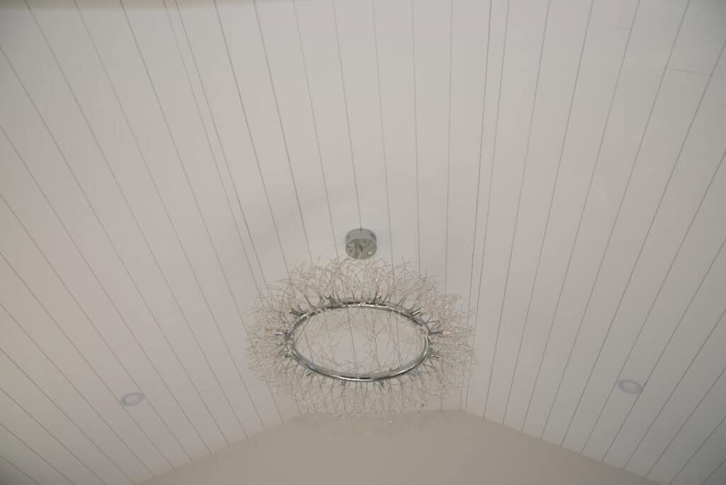 A modern chandelier with a circular metal frame and intricate wire design hangs from a white vaulted, paneled ceiling. Two recessed lights are visible on either side.