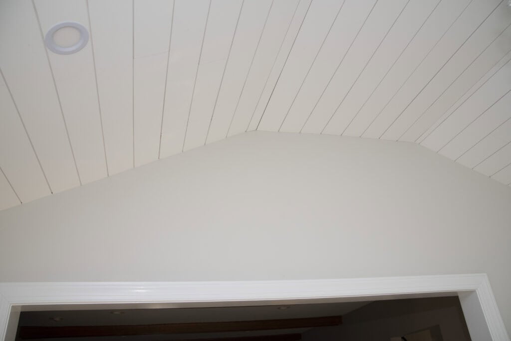 White, angled shiplap ceiling with recessed lighting above a doorway, featuring a slanted peak and smooth, light-colored walls.