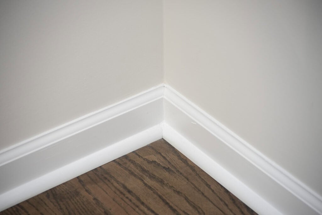 The image shows the corner of a room with light-colored walls, white baseboards, and a dark wood floor.