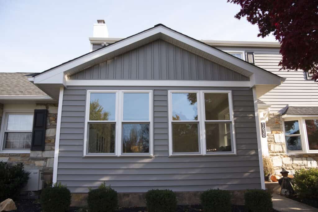 A close-up of a house exterior featuring gray vinyl siding, three large windows, stone accents, and white trim. Small shrubs and mulch line the base, and a red-leaved tree is partially visible on the right.