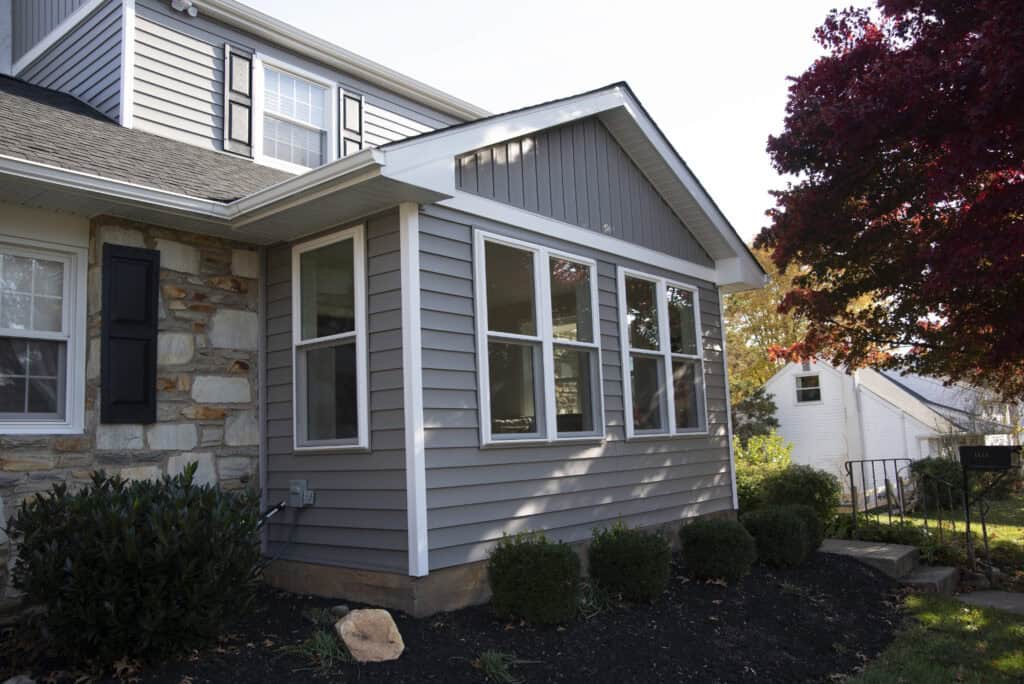 A gray house with white trim and large windows, featuring a stone accent wall and black shutters. Neat landscaping with bushes and a tree with red leaves.
