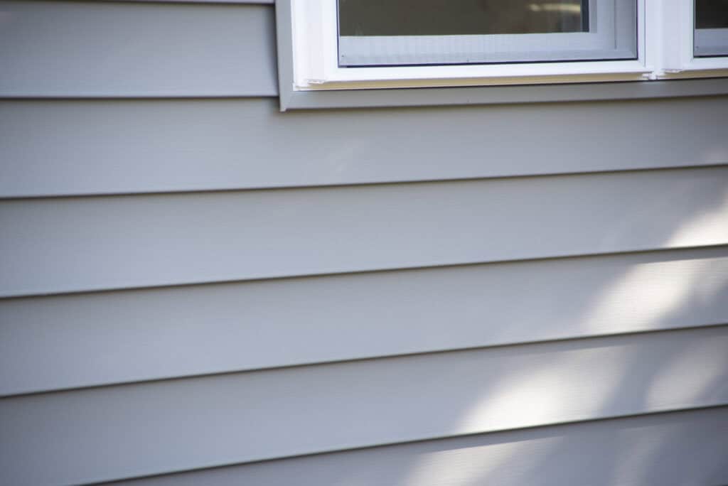 A close-up of light gray vinyl siding on a house with part of a white-framed window at the top. Sunlight and shadows fall across the siding, creating soft highlights and contrasts.