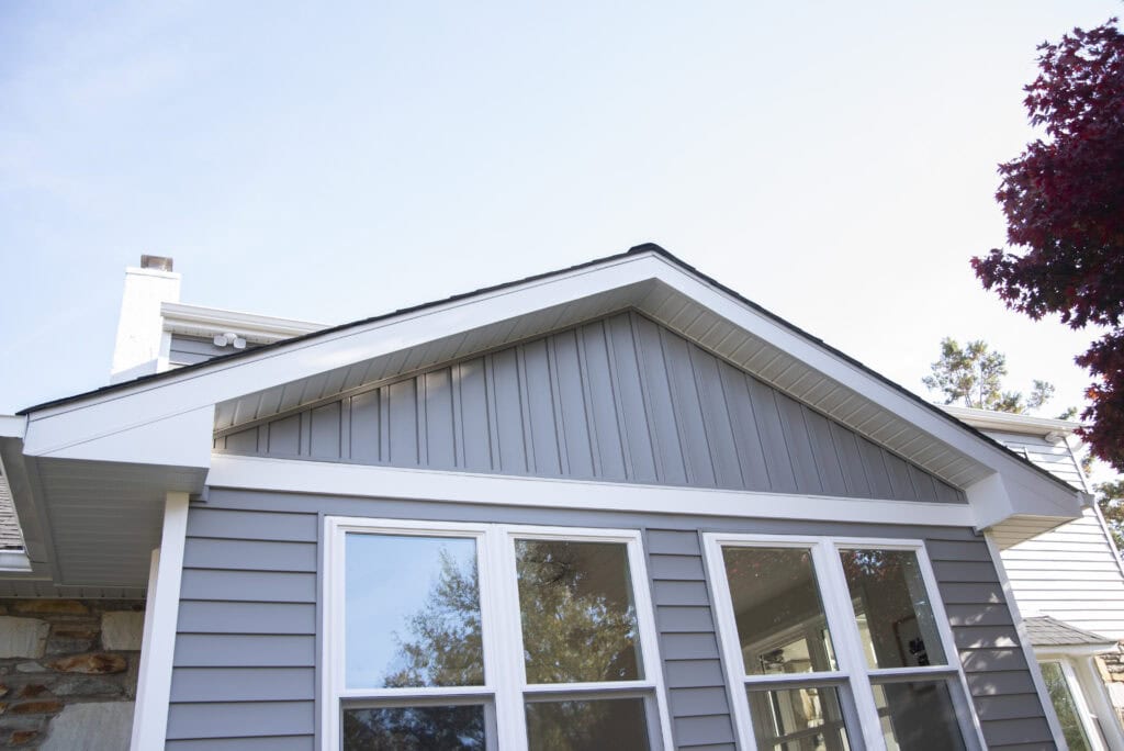 The image shows the exterior of a house with gray siding, white trim, and large windows. The roof is gabled, and there are trees and another house visible in the background.