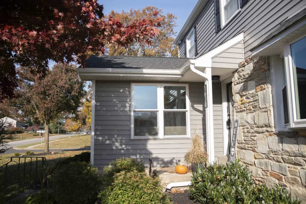 A gray house with stone accents and white trim is pictured on a sunny day. A pumpkin sits on the front walkway, surrounded by green shrubs, with colorful autumn trees in the background.