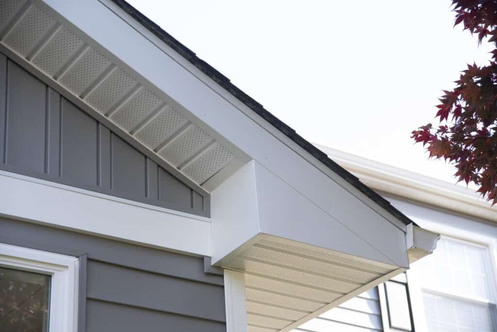 Close-up view of a house roofline featuring gray siding, white soffit, and fascia with vented panels beneath the eaves. A red-leaved tree and part of a neighboring house are visible on the right.