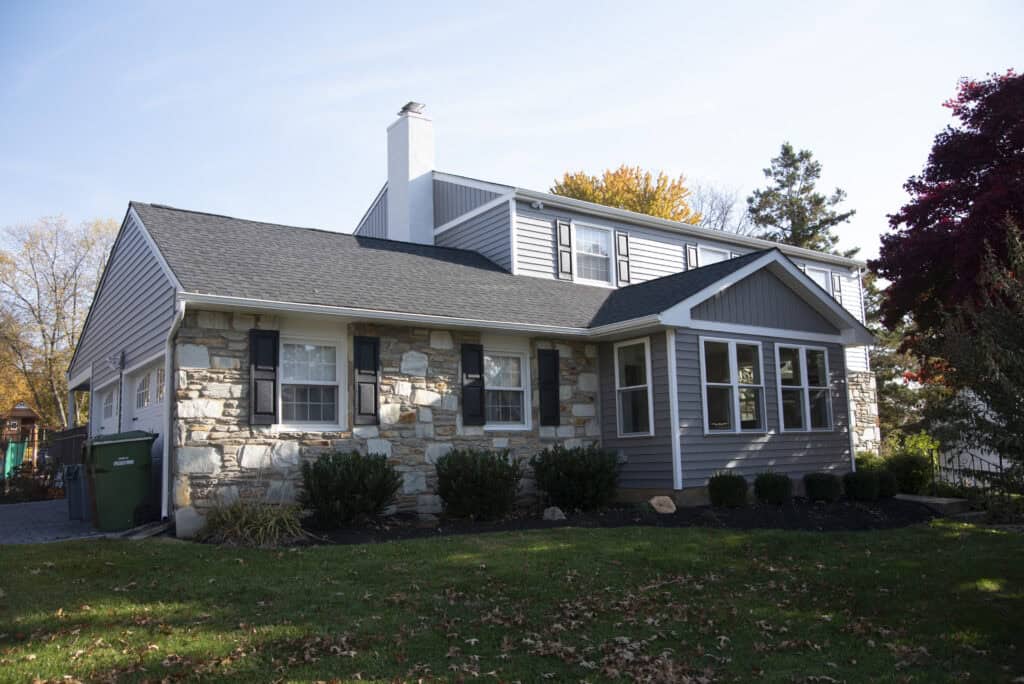A two-story suburban house with gray siding and stone accents, black shutters, and a white chimney, surrounded by bushes and grass on a sunny day in autumn.