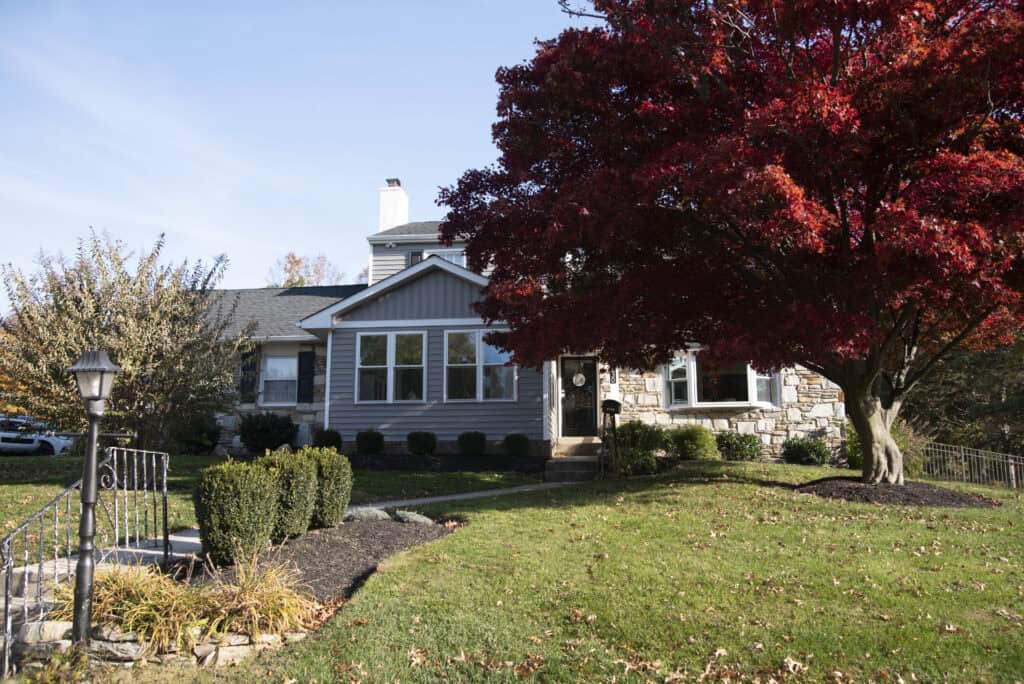 A gray house with large windows sits behind a well-kept lawn and a vibrant red-leaved tree. A black iron railing and a lamppost lead up the stone steps to the front entrance.