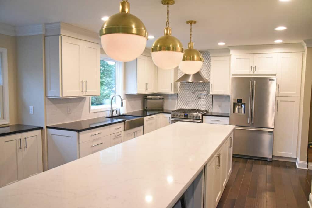 Modern kitchen with white cabinets, stainless steel appliances, a long white island, dark countertops, a patterned tile backsplash, and three large gold and white pendant lights hanging from the ceiling.