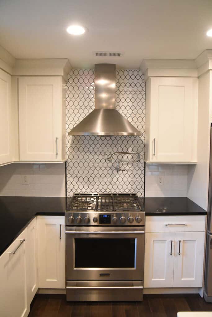 Modern kitchen with white cabinets, black countertops, a stainless steel gas stove and oven, and a stainless steel range hood. The backsplash features a white tile pattern with dark grout. The floor is dark wood.