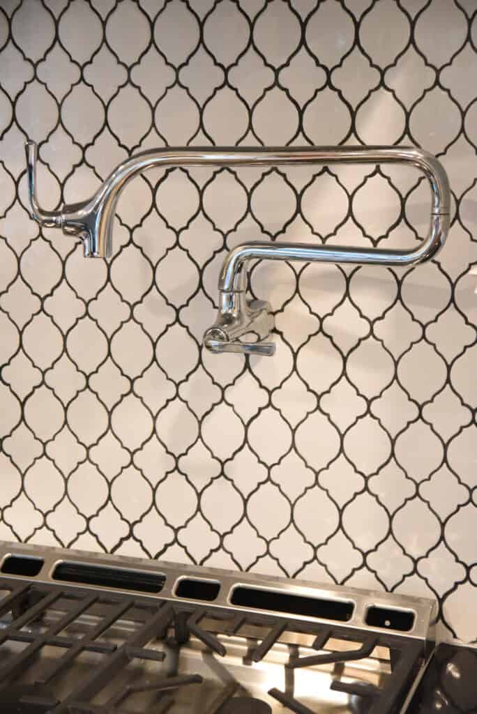 A shiny metal pot filler faucet mounted on a black and white patterned tile backsplash above a stove. The faucet has two jointed arms for flexibility.