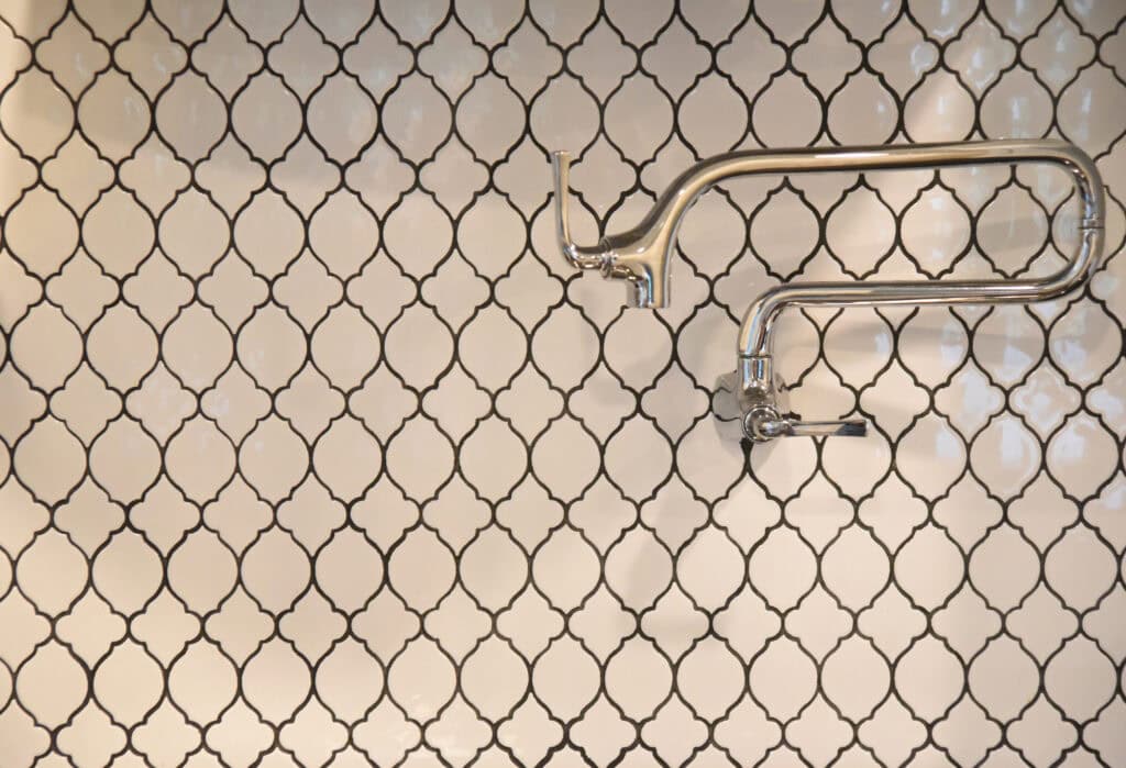 A section of wall with white and black arabesque patterned tiles, featuring a shiny silver faucet with two arms extending horizontally, one of which is missing its spout.
