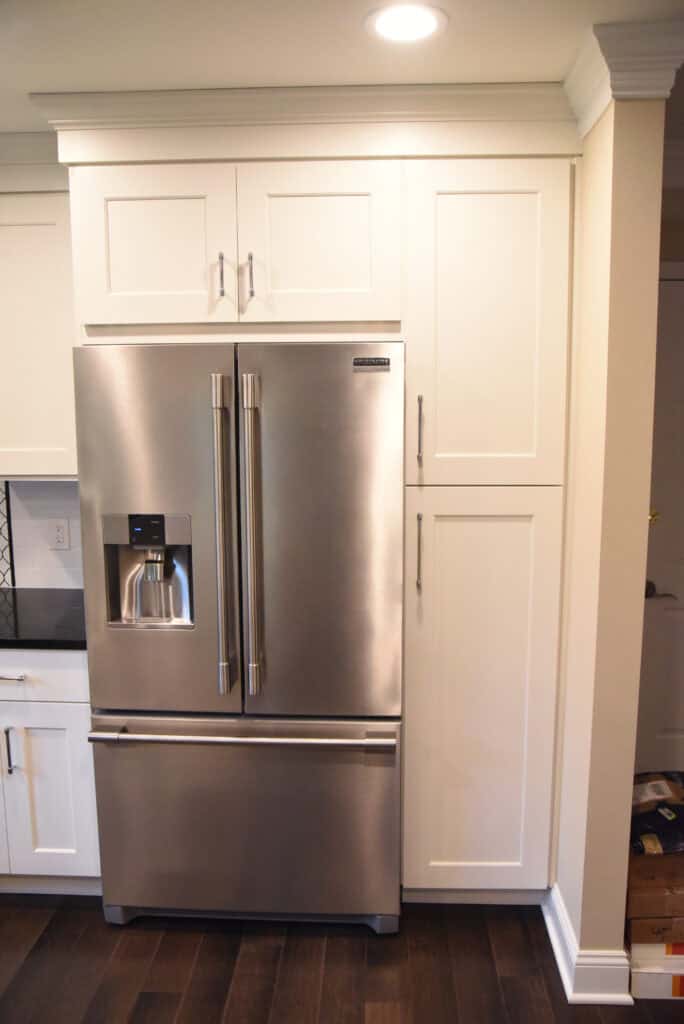 A stainless steel French door refrigerator with a water and ice dispenser, set within white cabinetry in a modern kitchen with dark wood flooring.