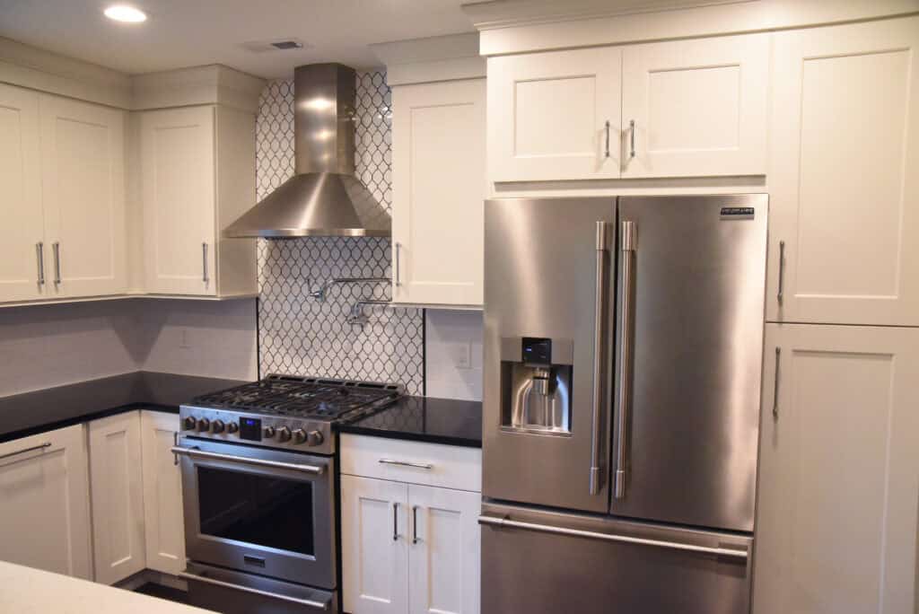 Modern kitchen with white cabinets, stainless steel appliances including a French door refrigerator and gas range, black countertops, and a patterned tile backsplash behind the stove.