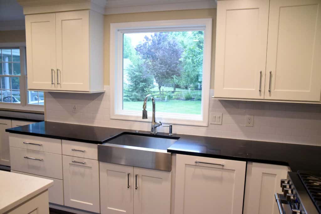 Modern kitchen with white cabinets, black countertops, a large stainless steel farmhouse sink, and a window above the sink showing a view of trees and greenery outside.