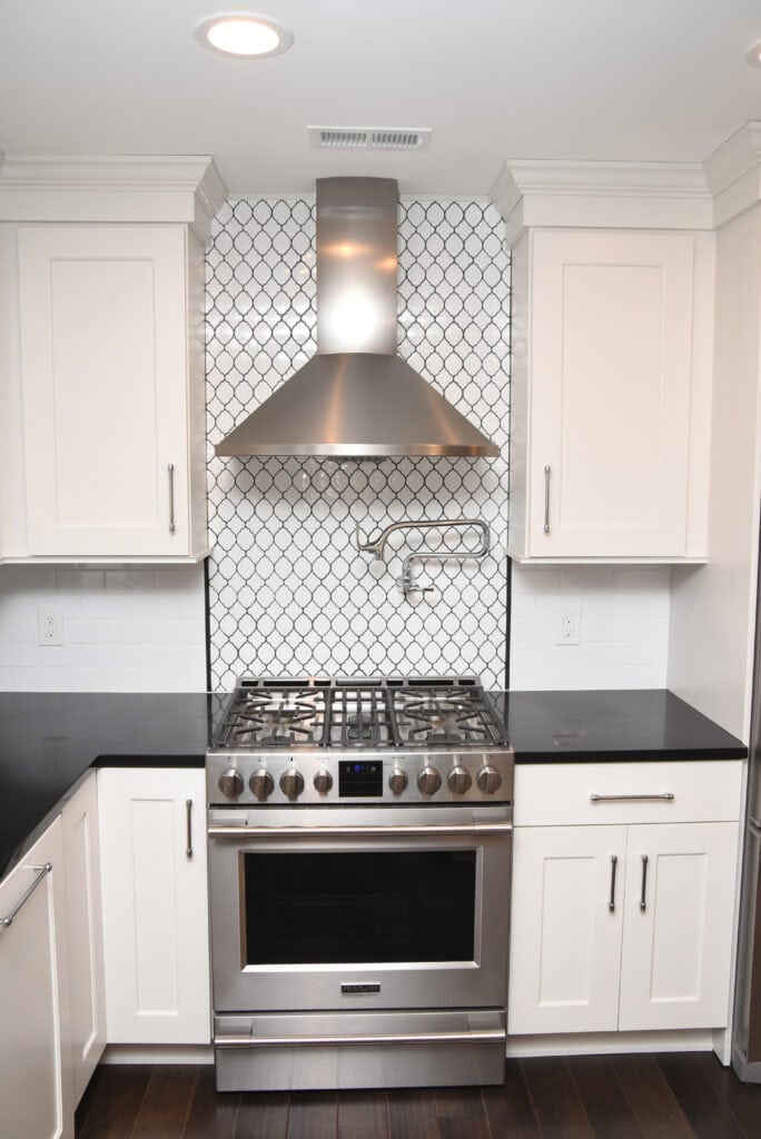 A modern kitchen with white cabinets, black countertops, a stainless steel gas stove and oven, a matching range hood, and a white backsplash with a black geometric pattern.