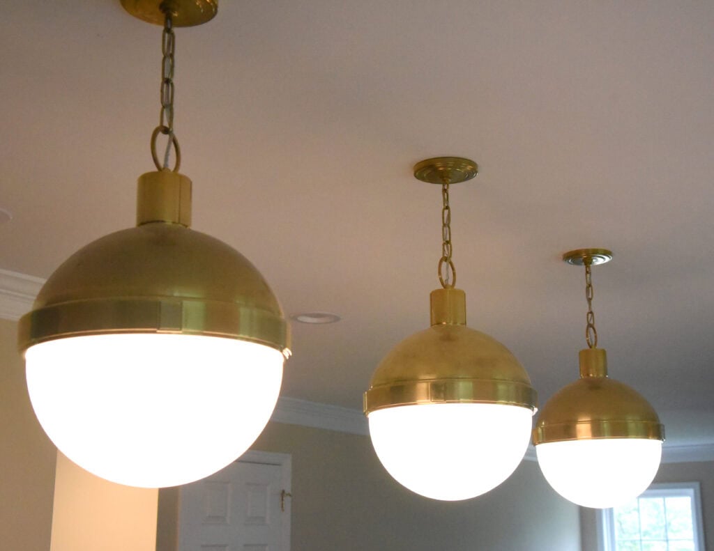 Three brass pendant lights with round white glass globes hang in a row from the ceiling in a well-lit room with beige walls, crown molding, and a window in the background.