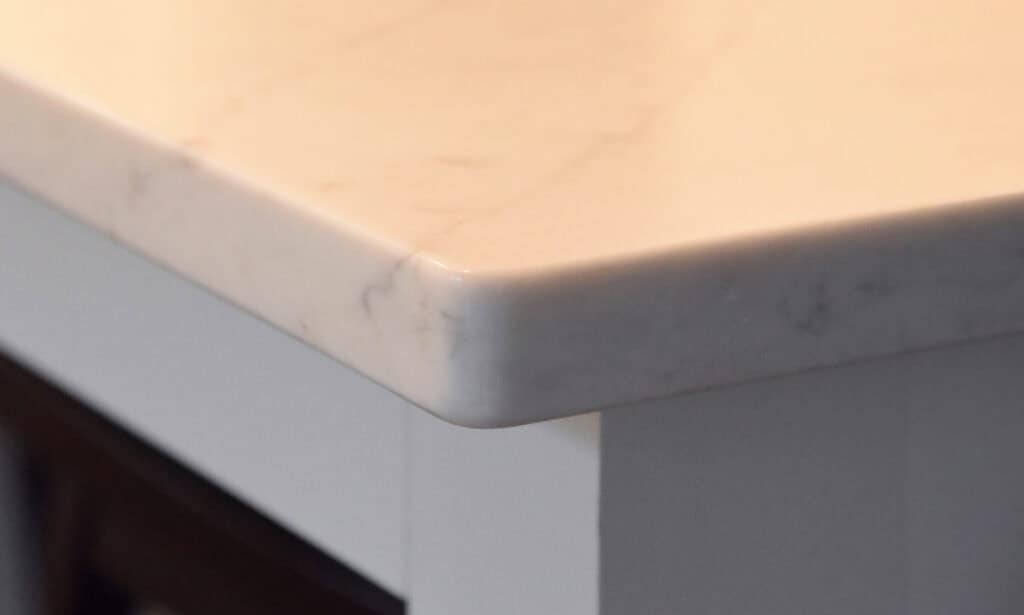 Close-up of a smooth, light-colored marble countertop with a rounded edge, showing subtle veining. The countertop sits atop a white cabinet.