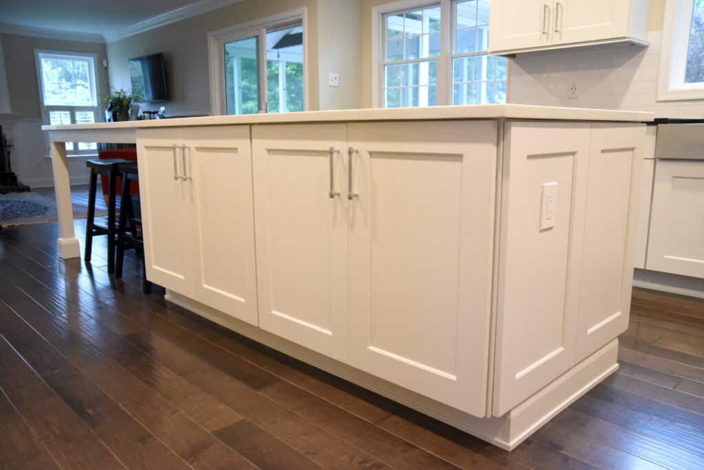 A modern kitchen island with white cabinets, silver handles, and a white countertop sits on dark hardwood floors in a bright, spacious kitchen with large windows in the background.