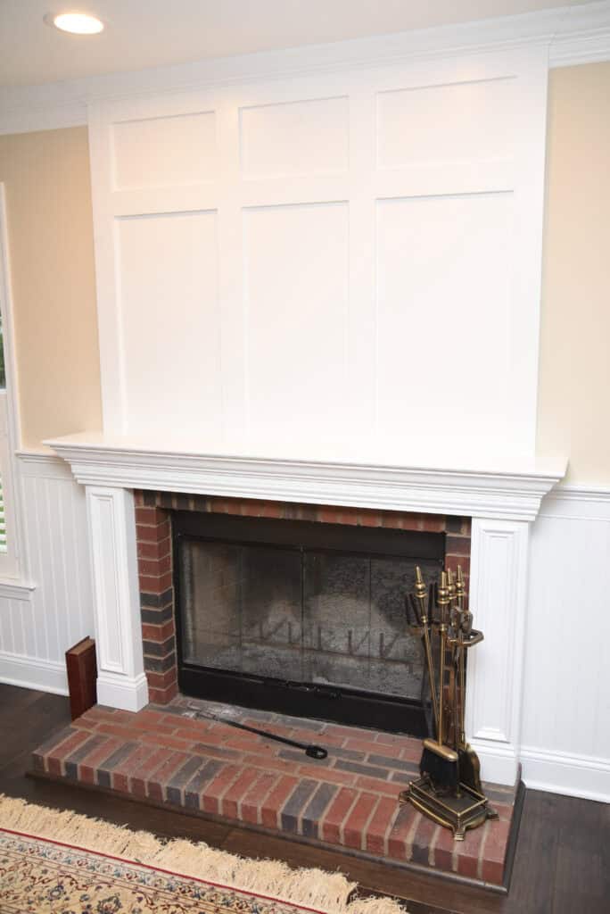 A brick fireplace with a white mantel and paneling above it; fireplace tools are on the right side of the hearth, and a patterned rug is partially visible in the foreground.