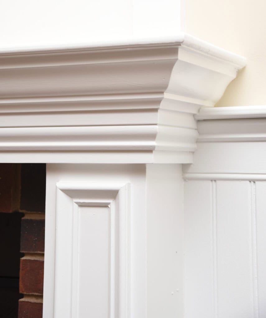 Close-up of a white, detailed wooden mantel with decorative moldings and trim, set against a cream wall with beadboard paneling and a glimpse of red brick inside a fireplace.