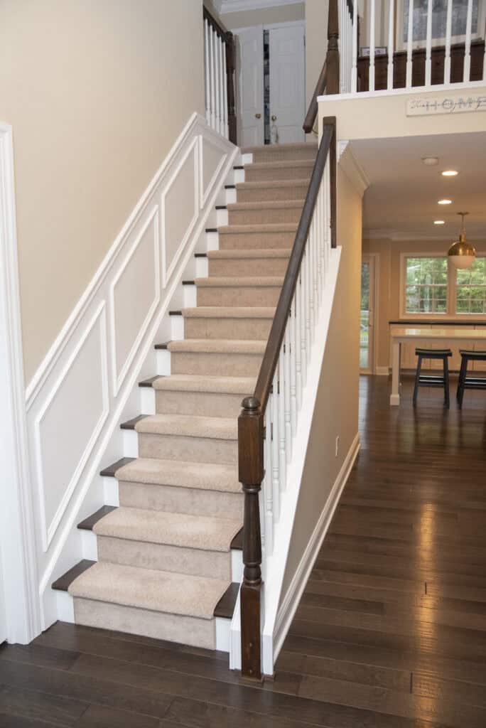 A carpeted staircase with white paneling and dark wood handrails in a modern home, leading to an upper floor; the kitchen and dining area with hardwood floors are visible to the right.