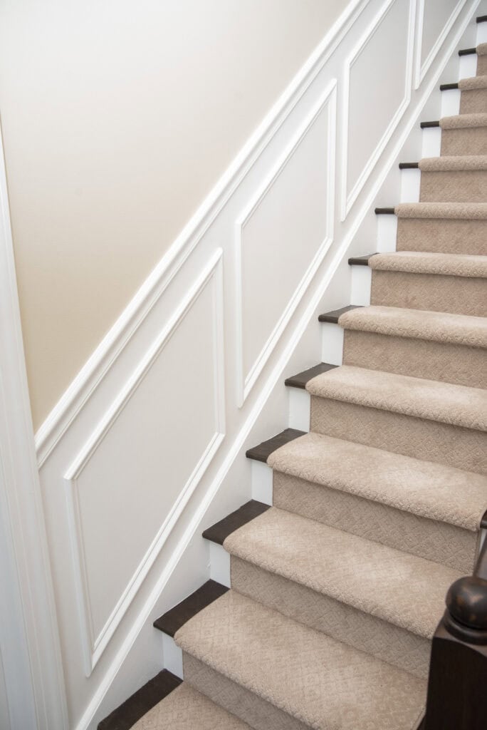 A carpeted staircase with light beige patterned carpet and dark wooden steps, bordered by white trim and decorative wall paneling on the left side of the stairway.