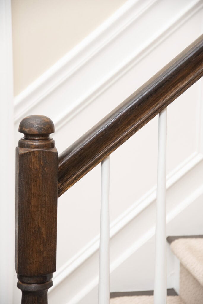 Close-up of a wooden staircase handrail with white balusters and a beige wall featuring white trim in the background. The steps are partially visible with light-colored carpeting.