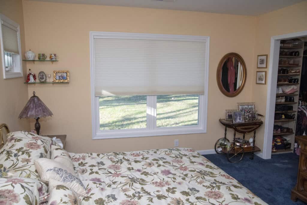 A bedroom with a floral bedspread, a window with a partially closed blind, peach-colored walls, a small wall shelf, a bedside lamp, a round mirror, and an open closet with shelves of shoes.