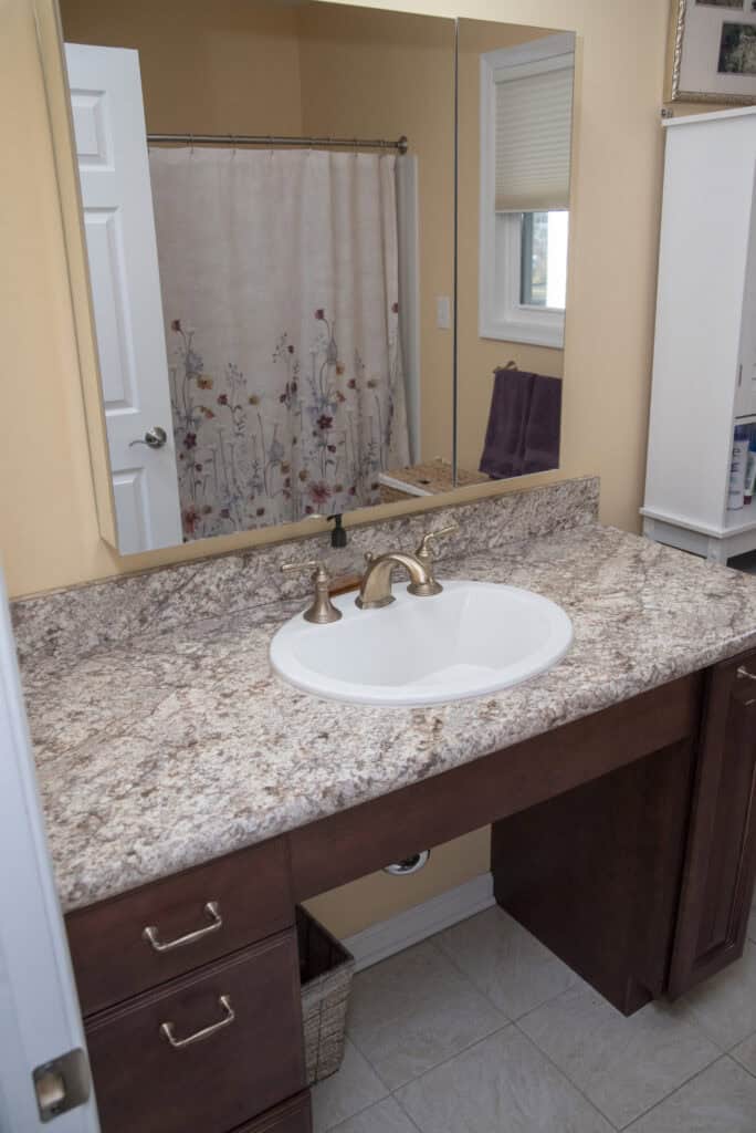 A bathroom vanity with a marble countertop, white sink, and brass faucet. There is a large mirror above the sink, and brown cabinets below. The background shows a shower with a floral curtain.