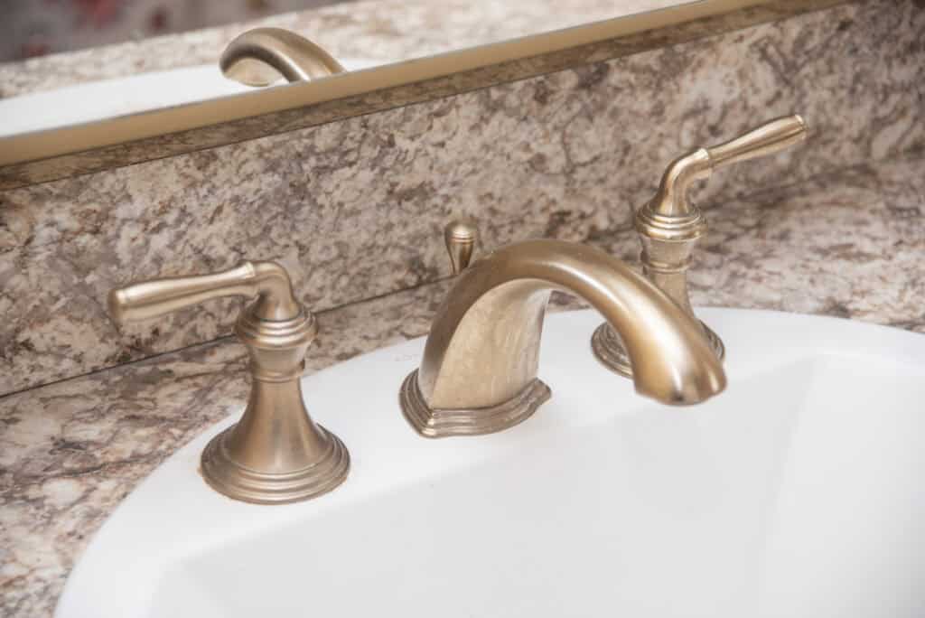 A close-up of a bathroom sink with a gold-colored faucet and two matching handles, set in a white basin with a marbled countertop and a mirror in the background.