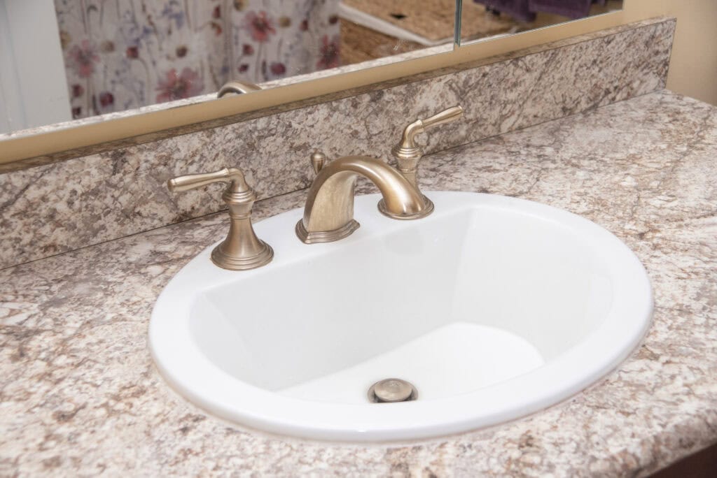Oval white bathroom sink with a marbled countertop, gold faucet, and two matching handles. A mirror is above the sink, and a floral shower curtain is visible in the background.