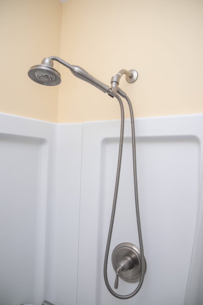 A modern shower fixture with a handheld showerhead attached to a flexible metal hose, mounted on a white shower wall with a beige painted upper section.