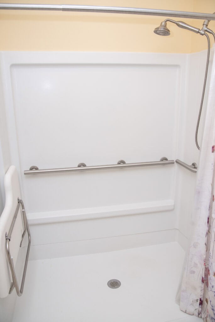 Accessible shower with a mounted showerhead, three horizontal grab bars on the back wall, a fold-down seat on the left, and a partially visible floral shower curtain on the right.