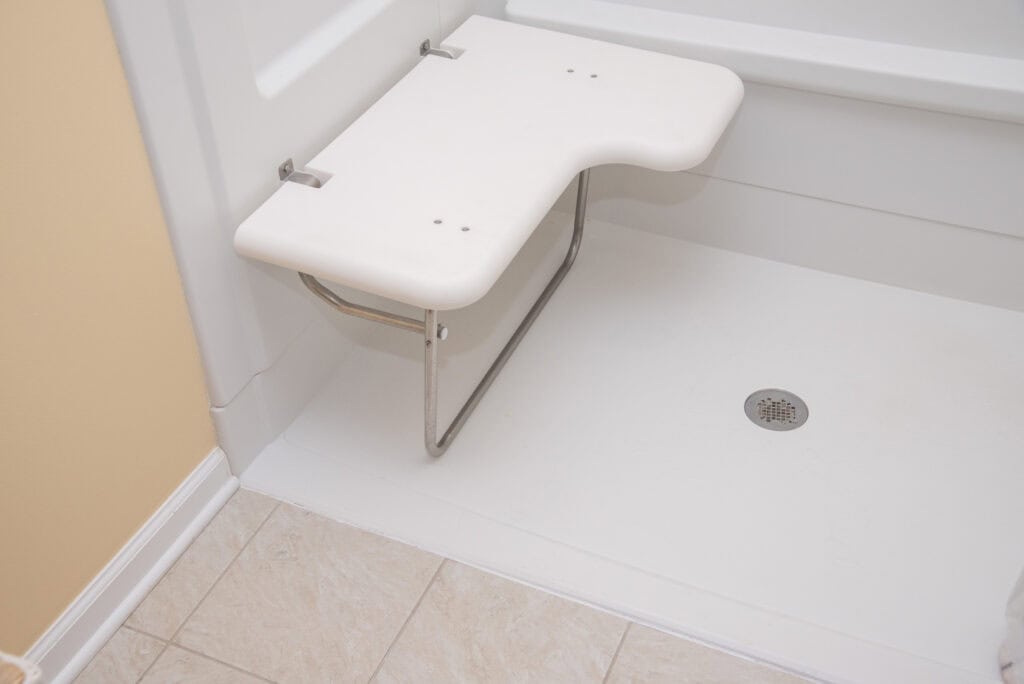 A white fold-down shower seat with metal legs is attached to the wall inside a walk-in shower, next to a tiled bathroom floor and beige wall.