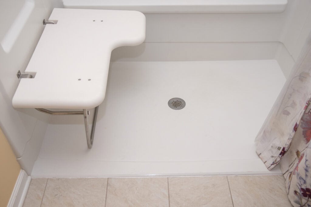 Accessible shower with a built-in white folding shower seat, beige tiled floor, white walls, and a floral-patterned shower curtain partially visible on the right side.