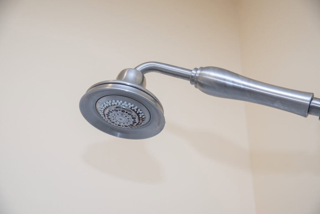 A close-up view of a silver showerhead with multiple spray settings, mounted on a slightly curved metal shower arm against a plain, off-white wall.