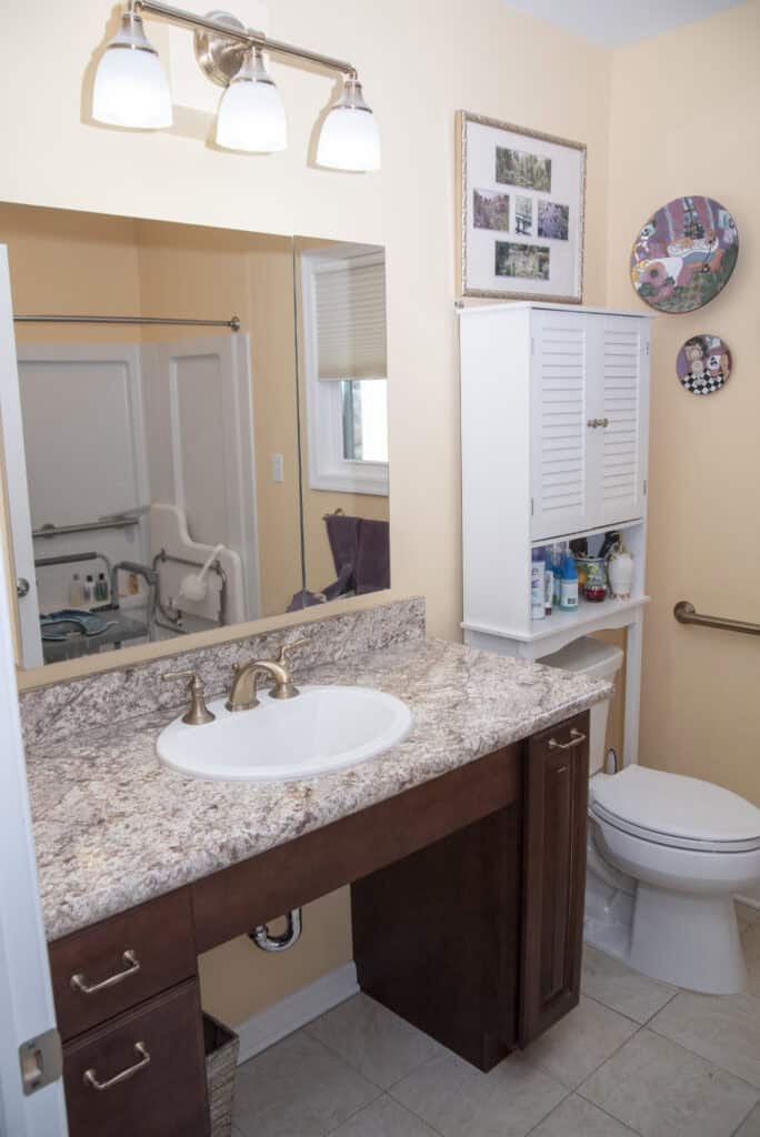 Accessible bathroom with a vanity sink, marble countertop, wall-mounted cabinet with toiletries, toilet with grab bars, walk-in shower with support bars, and framed art on walls. The room has beige walls and tiled floor.