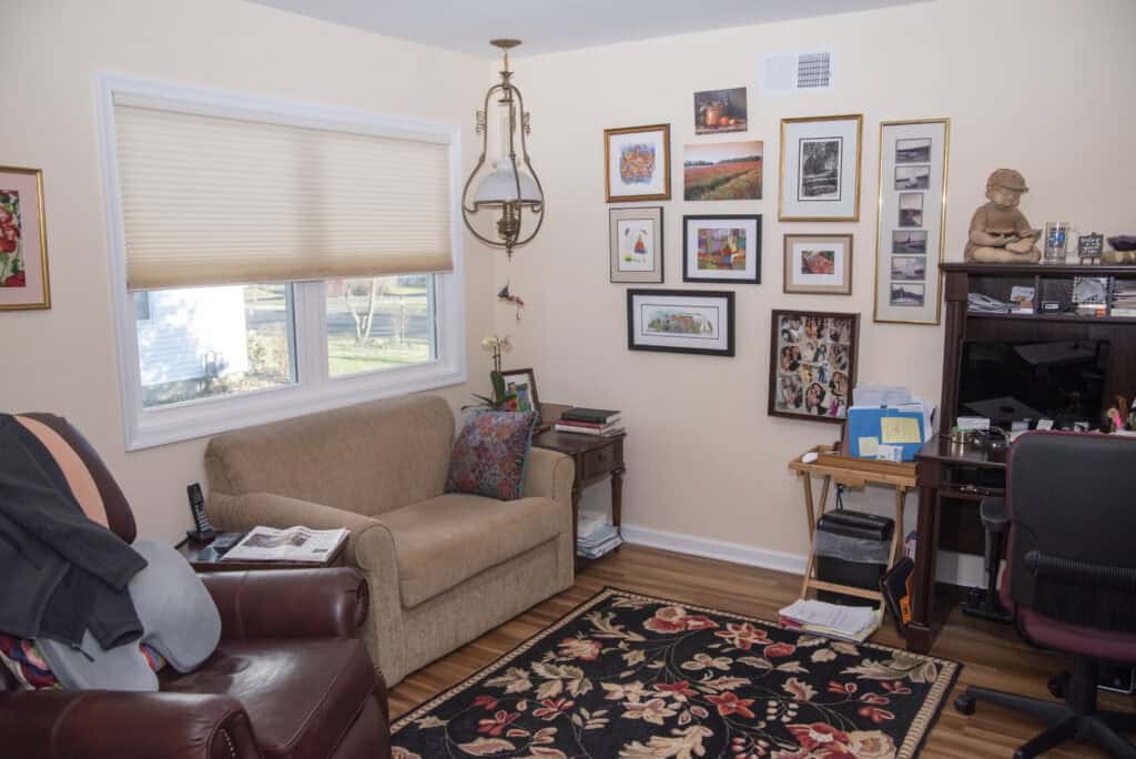 Cozy home office with beige walls, two armchairs, a patterned rug, wall art gallery, small desk with computer, lamp, and window with shades, decorated with books, photos, and a statue.