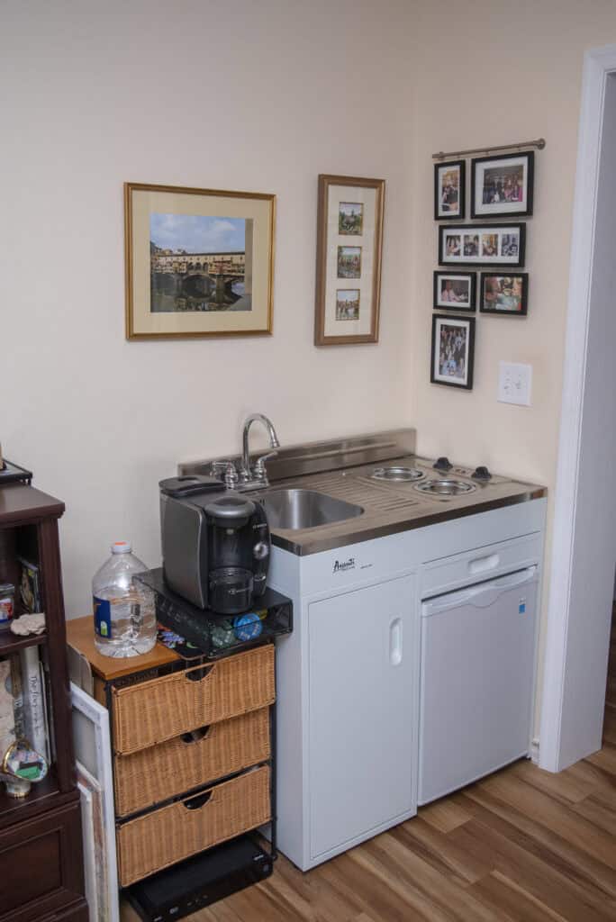 A compact kitchenette with a sink, two electric burners, a small fridge, and a black coffee maker. Wicker drawers and a water jug are beside it. Framed photos and art decorate the beige wall above.