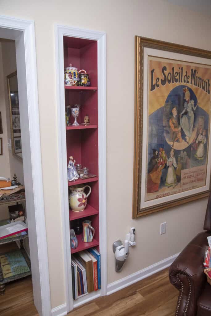A narrow pink shelving unit in a wall holds decorative teapots, cups, and figurines. Below are books and binders. A framed vintage poster hangs nearby, with a wall-mounted device and chair visible on a wood floor.